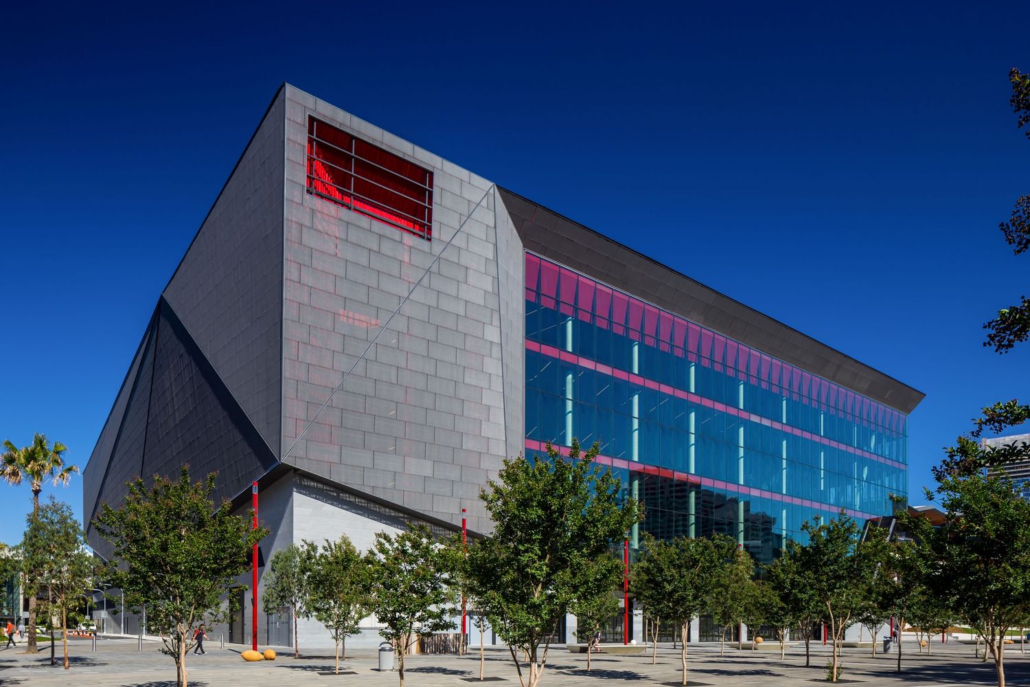 Modern convention center with angular gray panel facade, red accent window and blue glass wall, framed by trees under a clear sky.