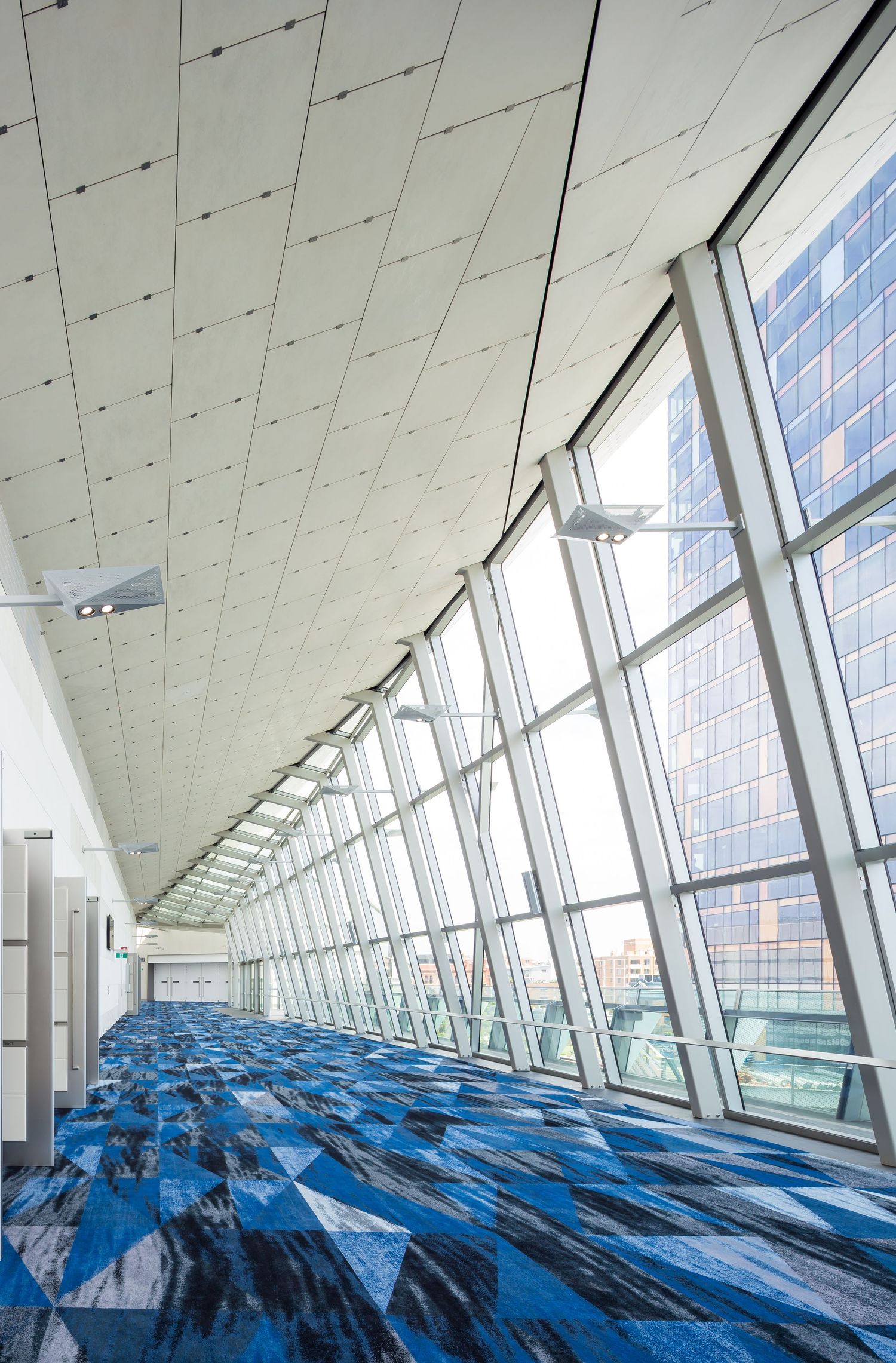 Long convention centre corridor with a patterned blue carpet, angled glass wall and city views.