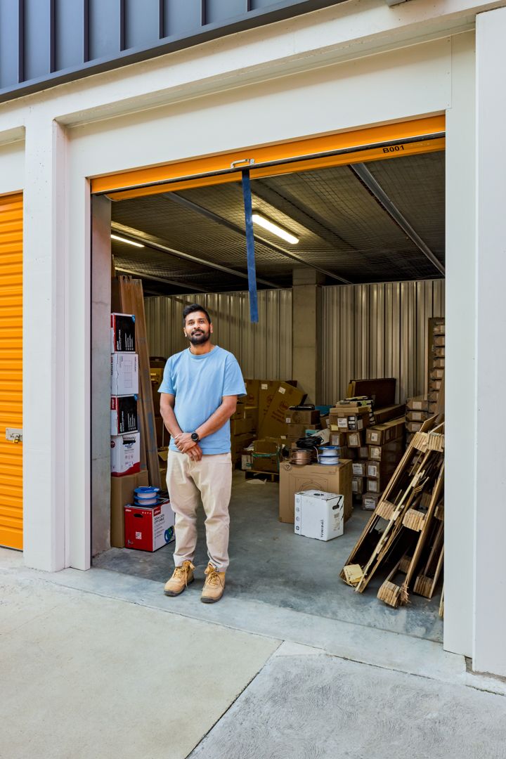 A bearded man in a light blue shirt and khaki pants stands in the doorway of an open storage unit filled with boxes and timber.