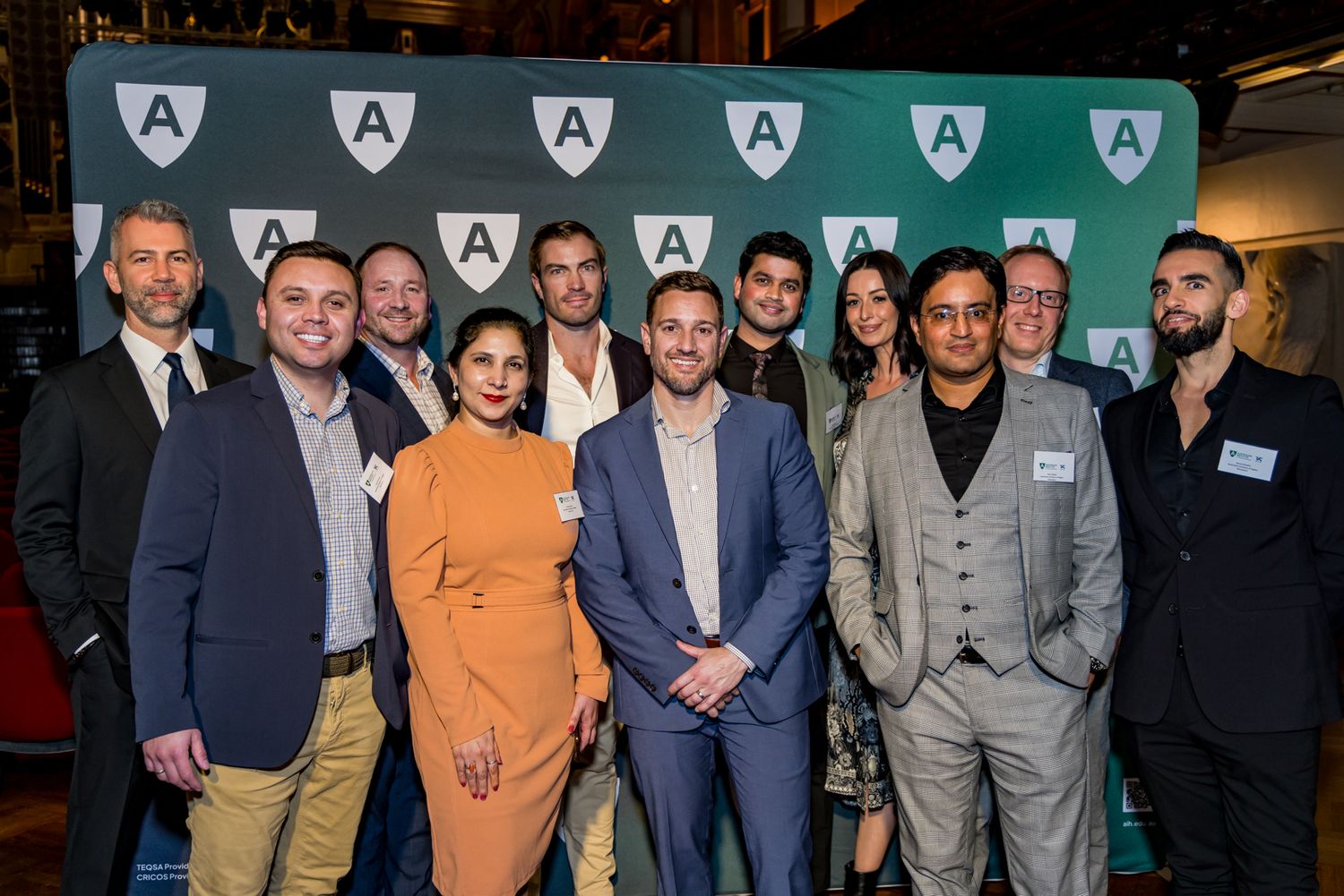 A group of men and women in business attire pose together at a networking event in front of a green backdrop with repeating logos.