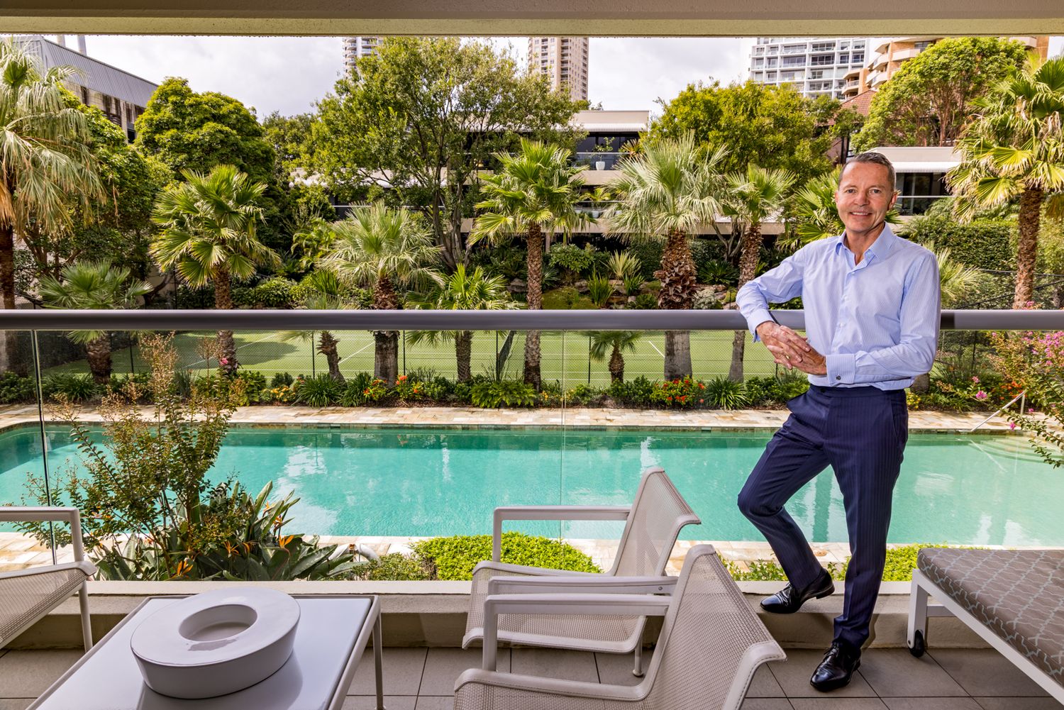 Smiling man in a blue shirt and navy trousers stands on a balcony overlooking a pool, palm trees and apartment buildings.