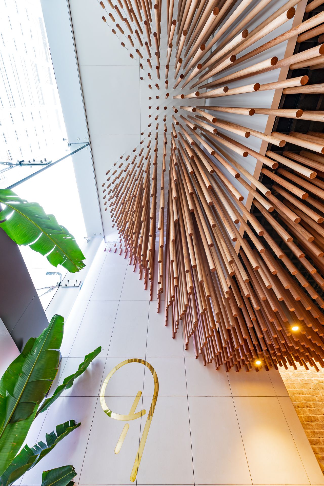 Radiating timber dowel sculpture on a lobby ceiling with tropical plants and a gold number nine sign on the floor.