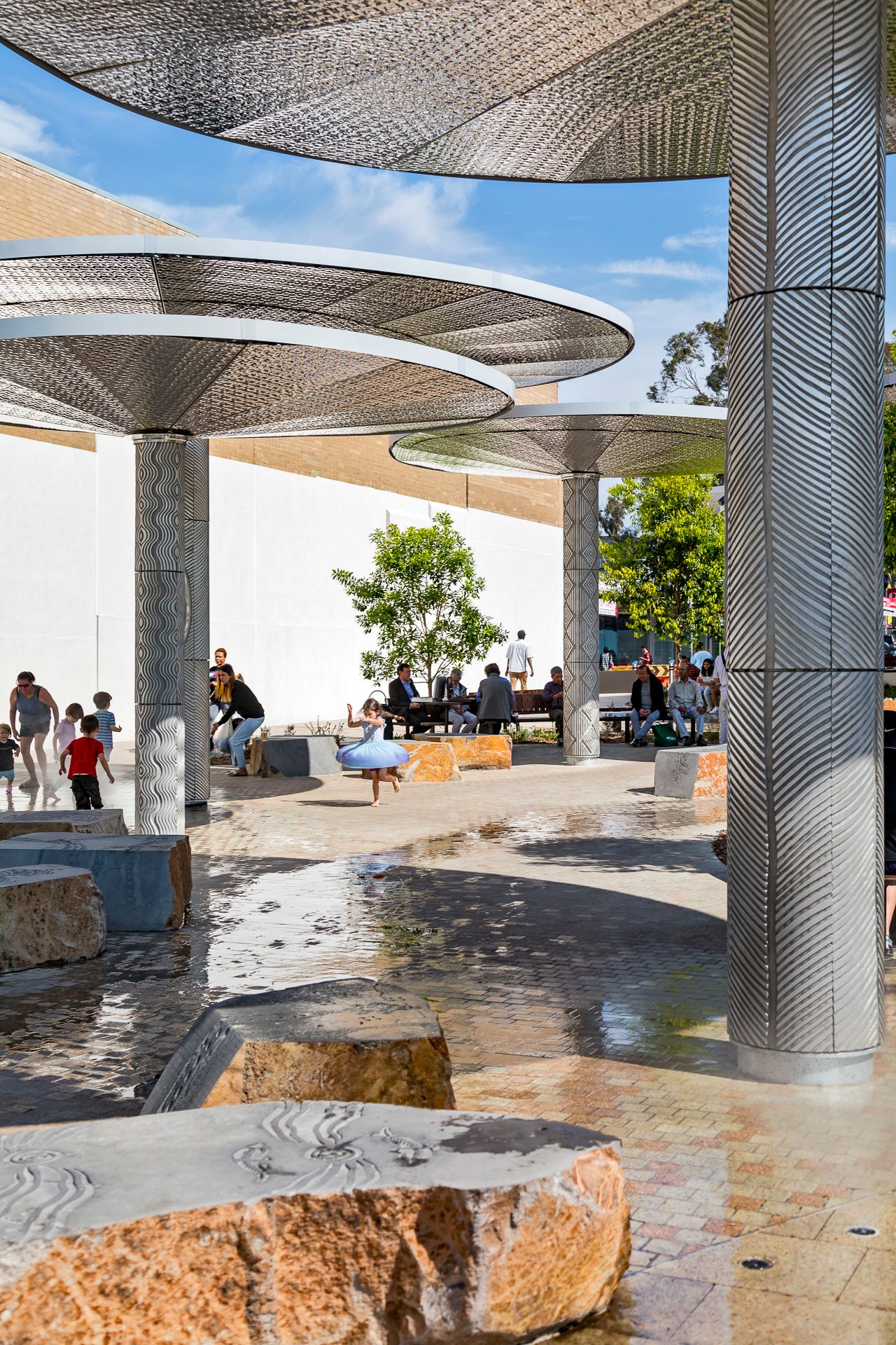 Children play and visitors sit under patterned metal umbrella shade structures above carved sandstone seats on a wet paved town square.