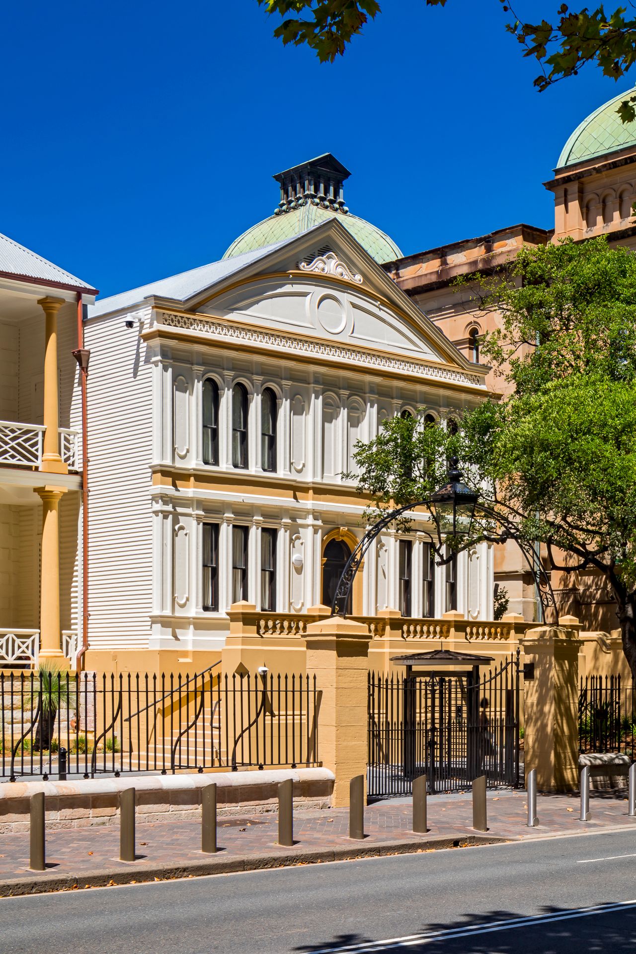 Historic two-story building with neoclassical facade, arched windows, green copper dome and wrought-iron fence under a clear blue sky.