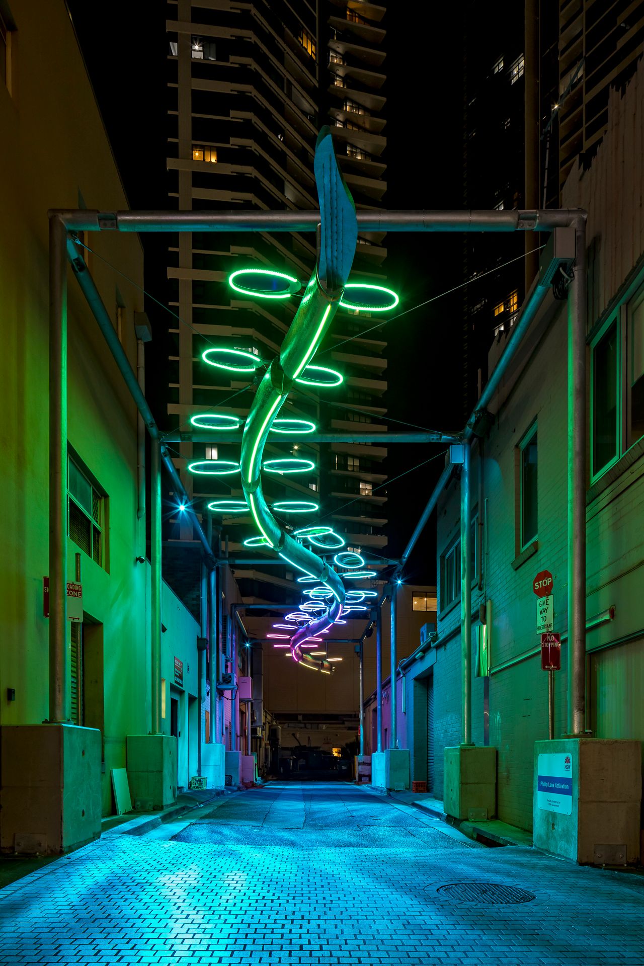 Green and pink neon rings spiral over a teal-lit alley with brick walls and a skyscraper in the background.