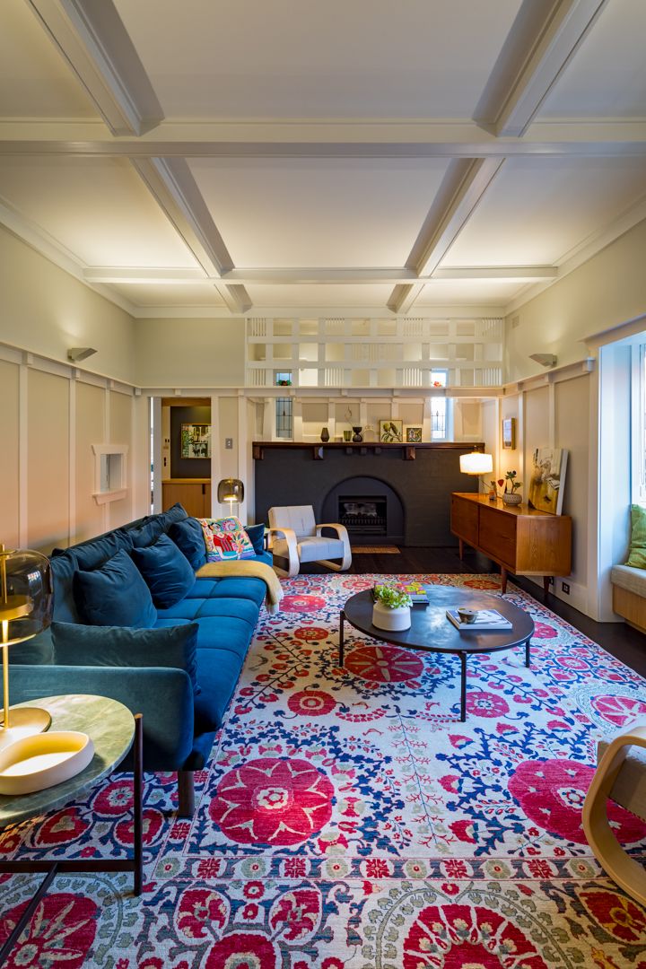 Elegant living room with coffered ceiling, blue velvet sofa, bold floral rug, dark fireplace and warm lighting.