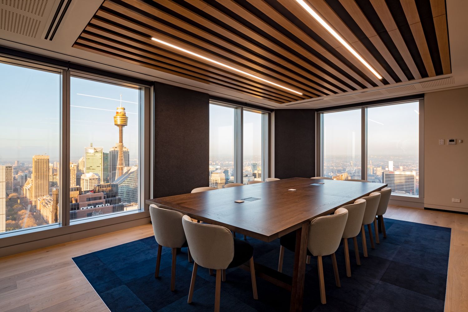 Modern boardroom with a wooden table, cream chairs and navy rug, framed by floor-to-ceiling windows with Sydney skyline and tower views.