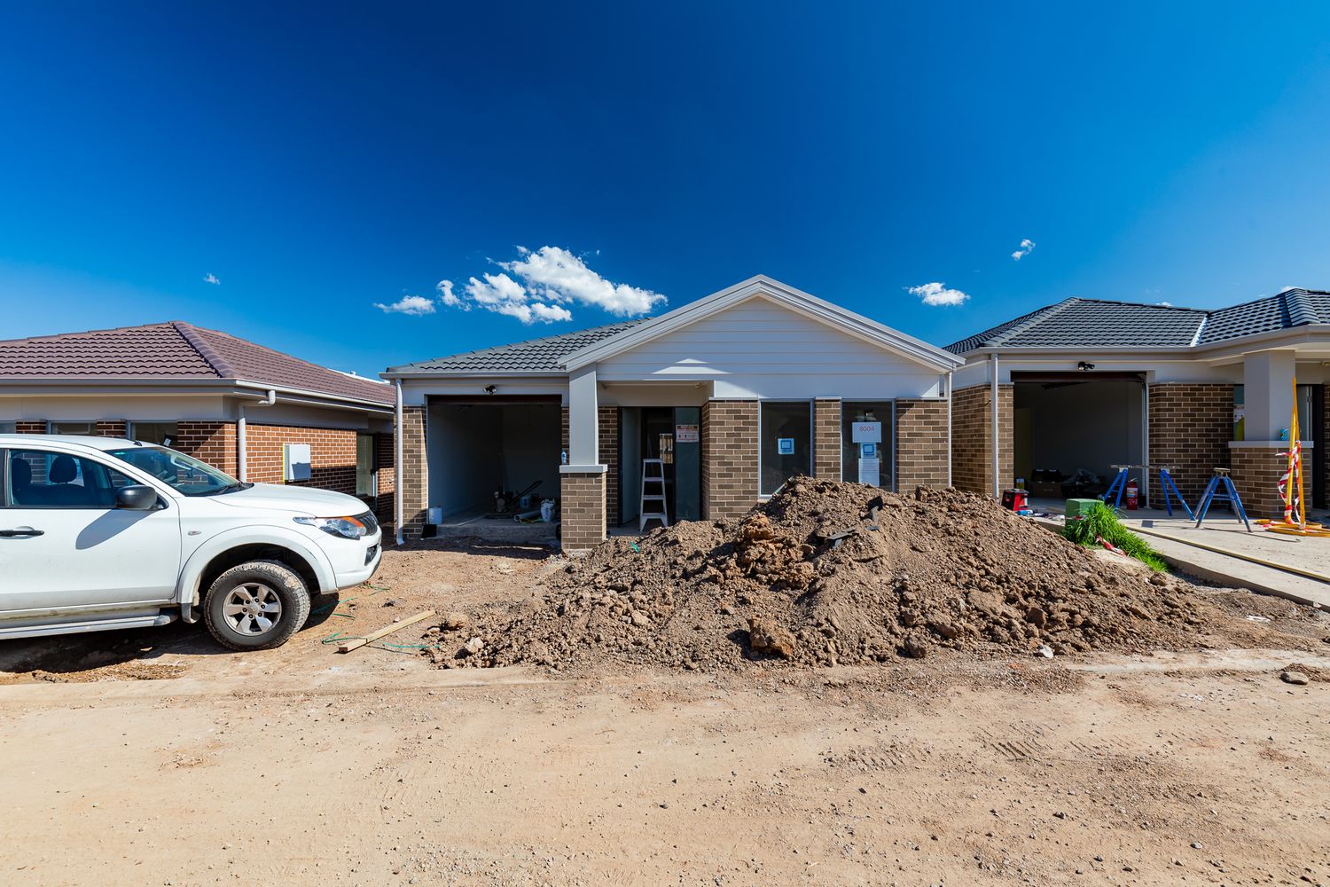 Brick home with covered porch nearing completion on a building site, with a pile of excavated soil and a white ute parked beside it.