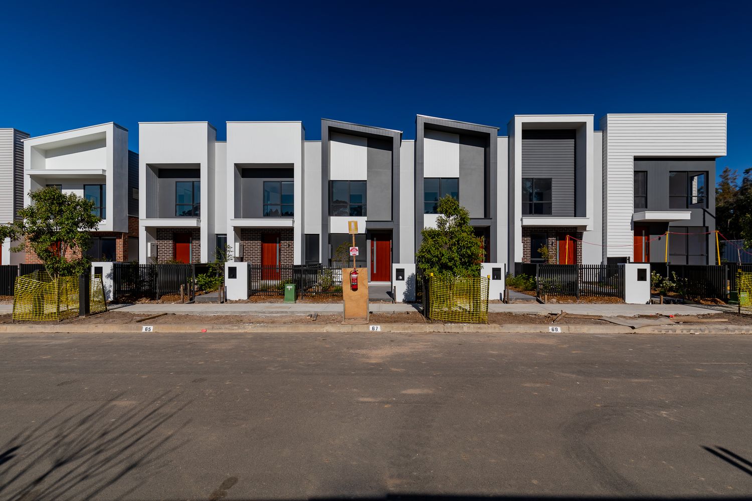 Row of modern white and render townhouses with red front doors, black fencing and young trees along a quiet suburban street.