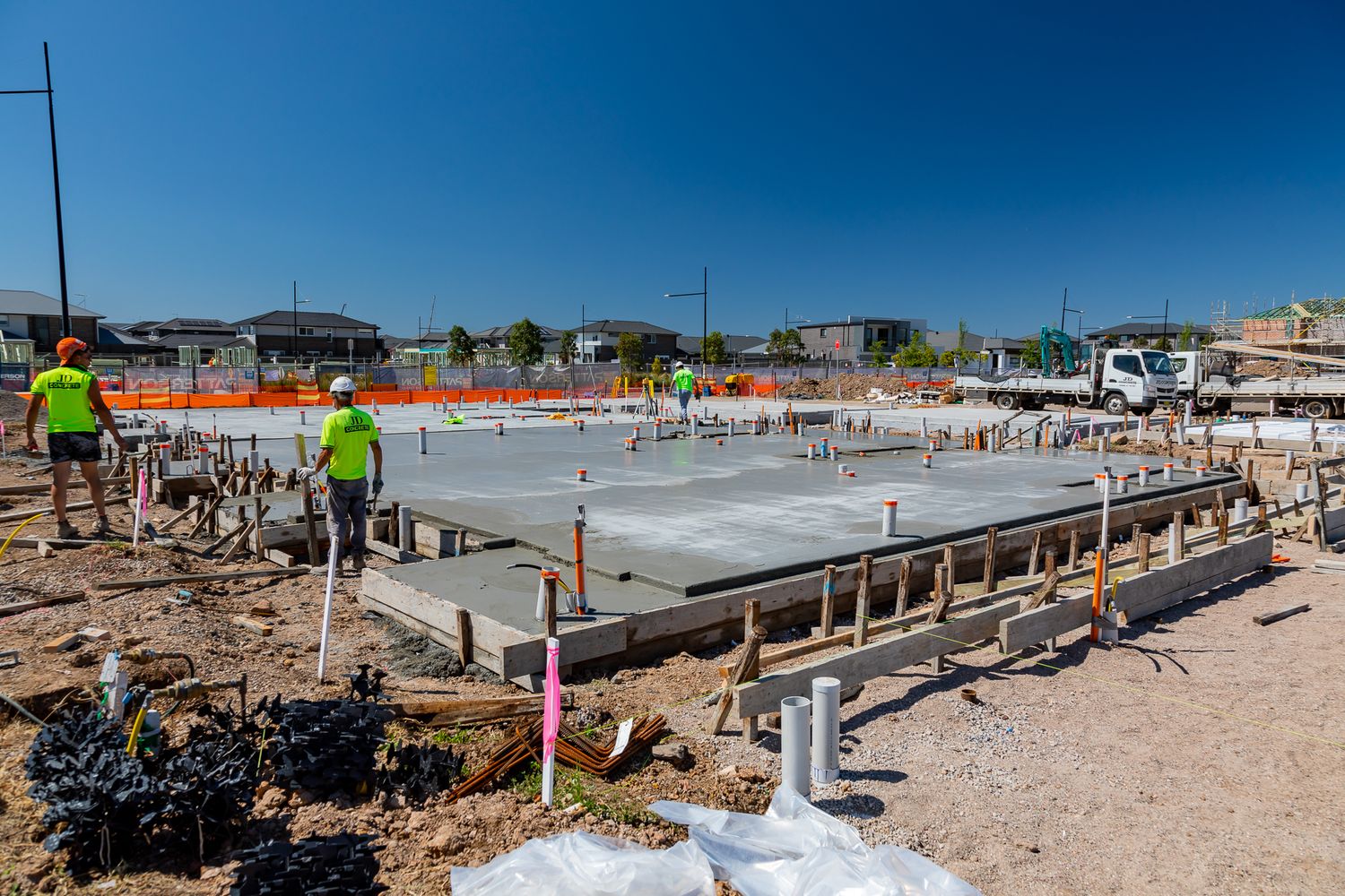 Workers in hi-vis shirts and hard hats prepare a concrete slab with timber formwork and steel reinforcement at a new housing site.