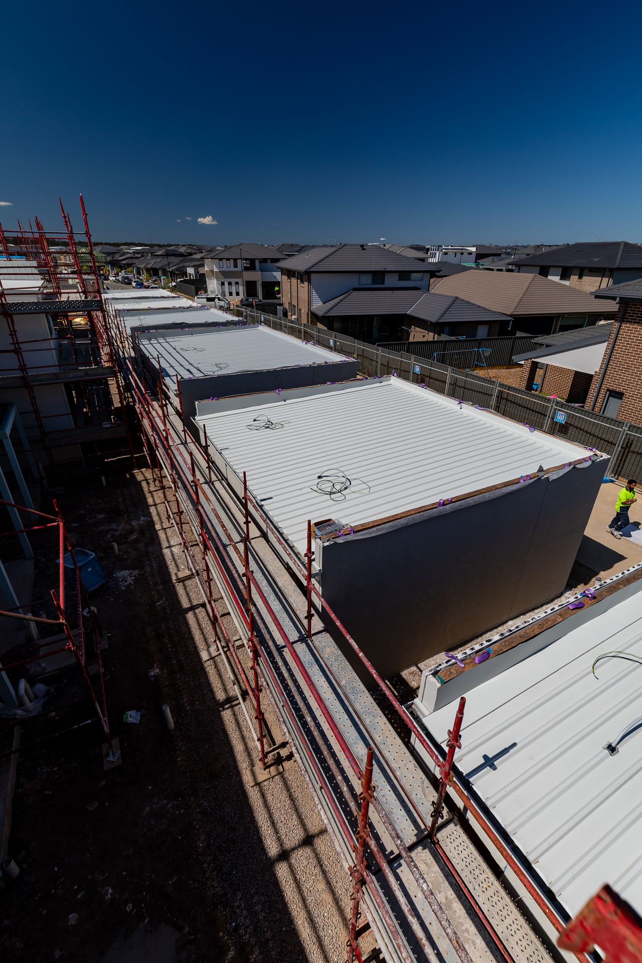 High angle view of townhouse roofs with metal sheeting and red scaffolding, overlooking a suburban housing estate on a clear day.
