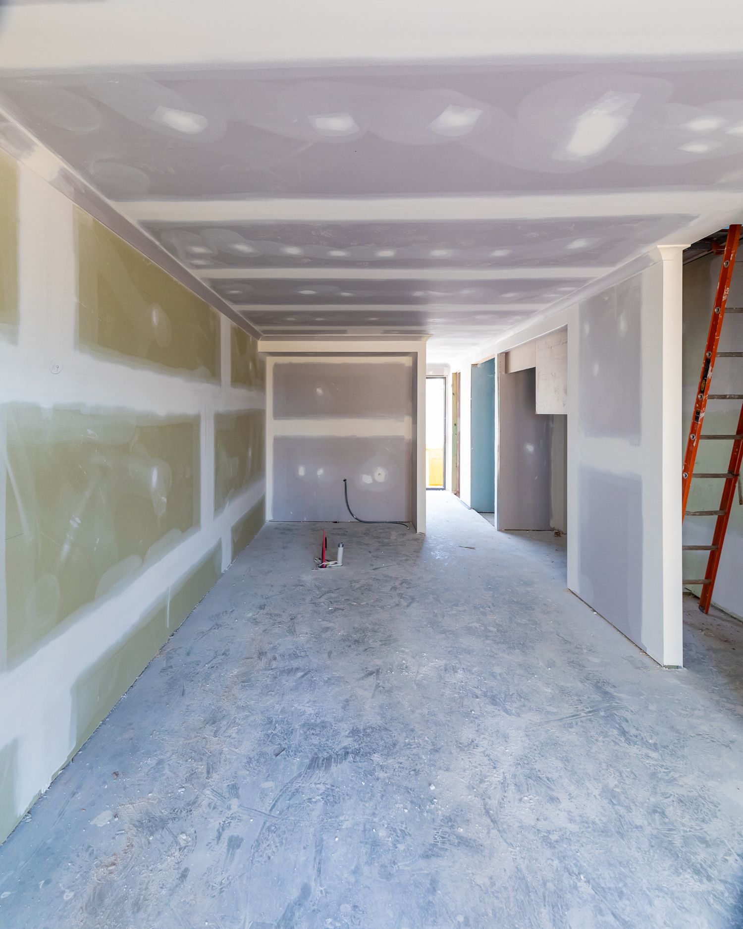 Interior of an unfinished home with plasterboard walls and ceiling, bare concrete floor, and a red ladder at the side.
