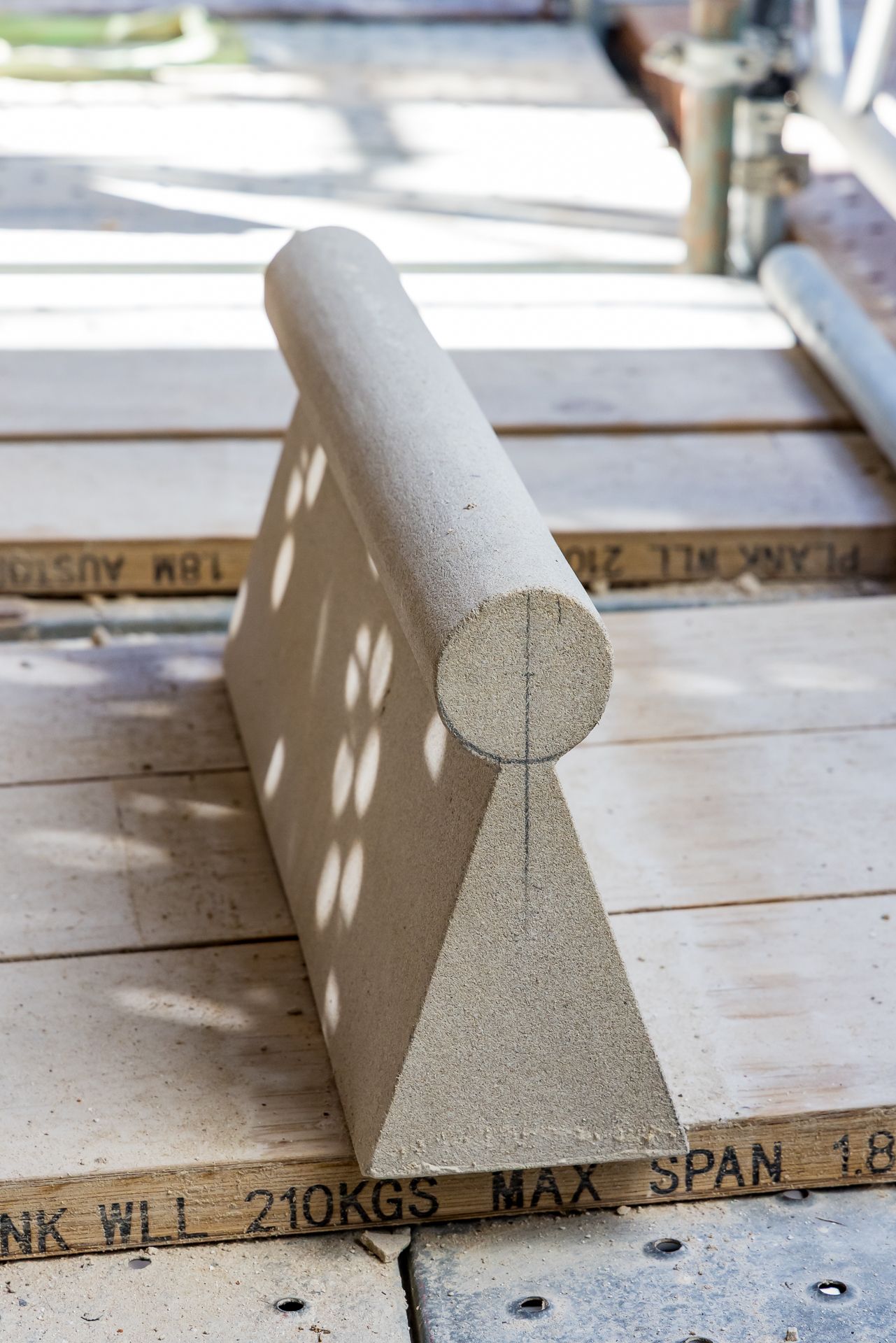 Sculpted stone ridge piece with a rounded top and triangular base resting on scaffold planks in sunlight.
