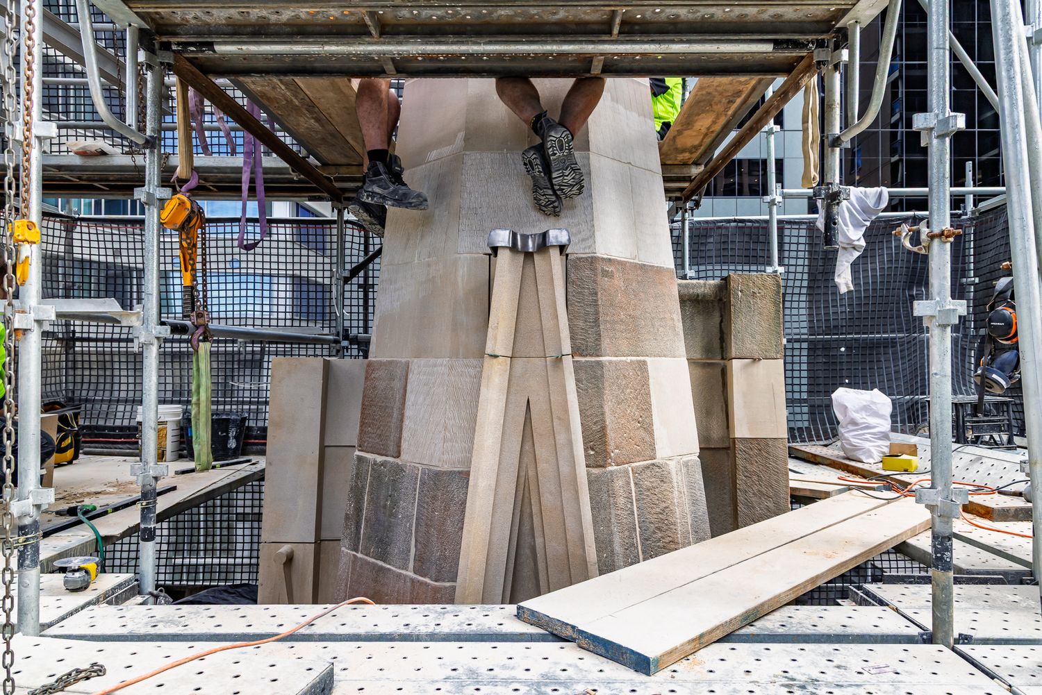 Stonemasons' boots dangle from scaffolding above a newly carved sandstone buttress base during historic building restoration.