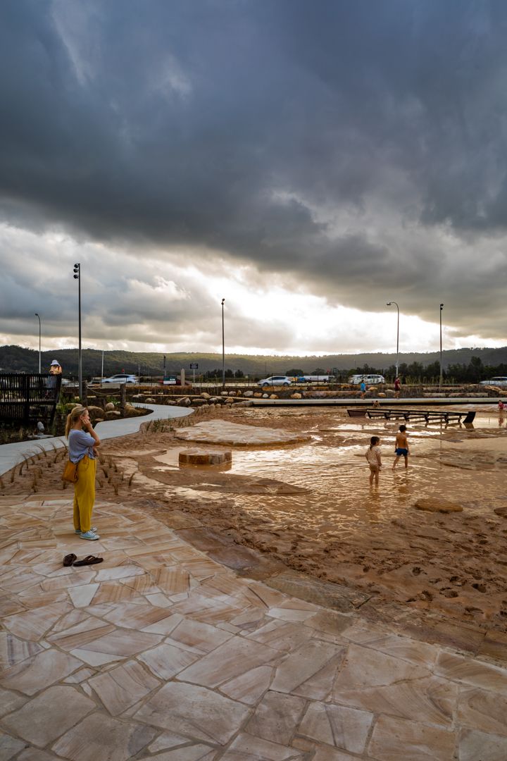 Woman in a yellow dress stands on stone paving as children play in shallow water at Gosford Leagues Club Park under stormy clouds.
