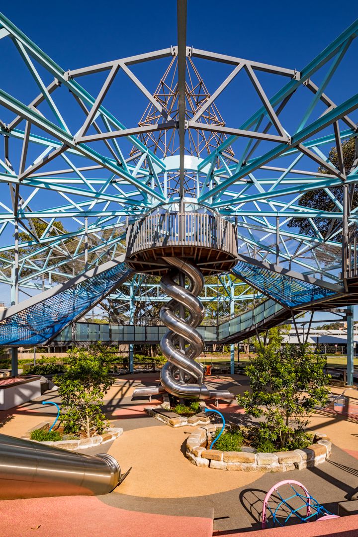 Playground with a tall steel truss structure, spiral metal tube slide and elevated walkways above sandy ground and landscaped planters.