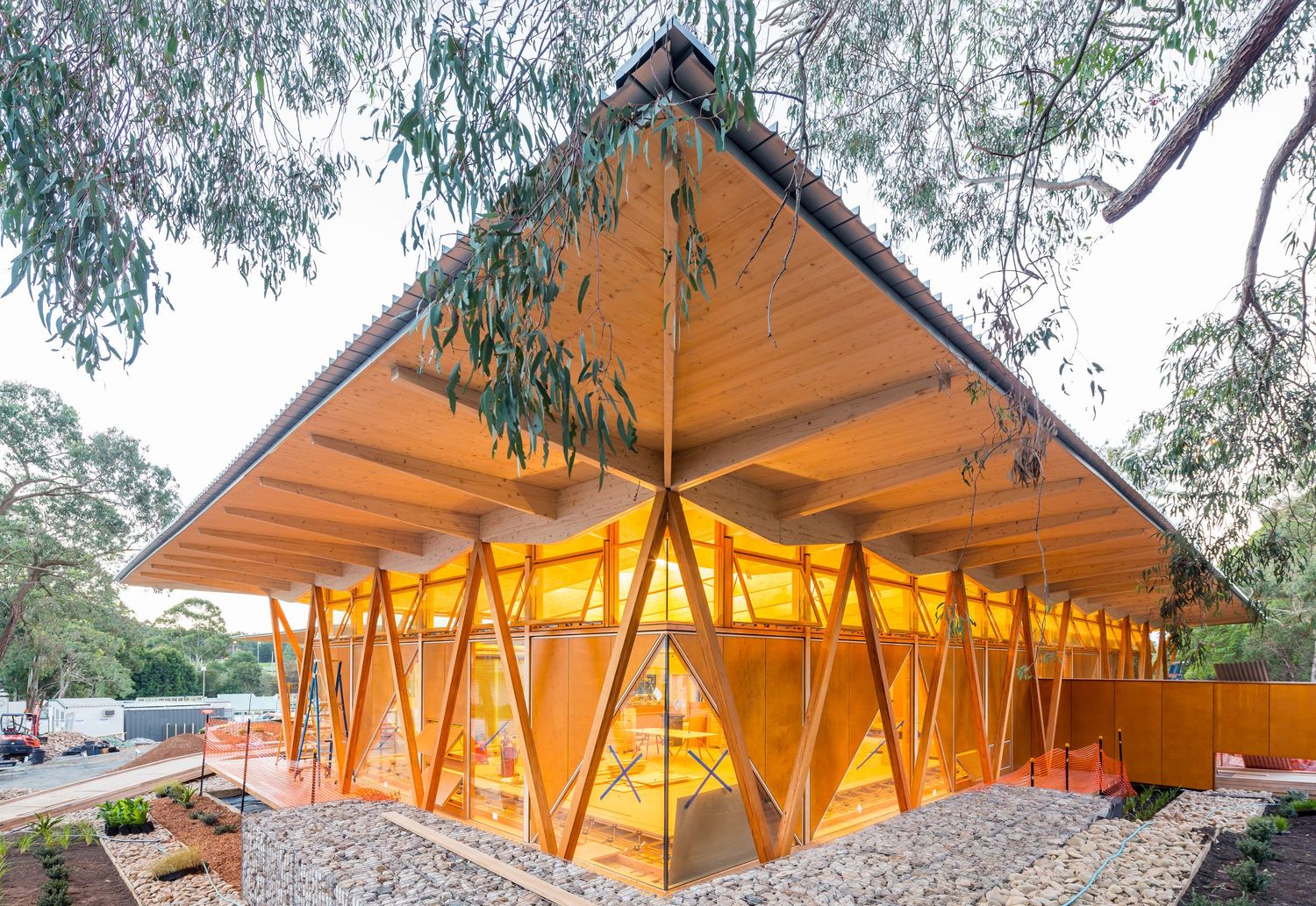 Timber-framed pavilion with angled wooden columns and a pitched roof glowing amber beneath gum trees and a gravel path.
