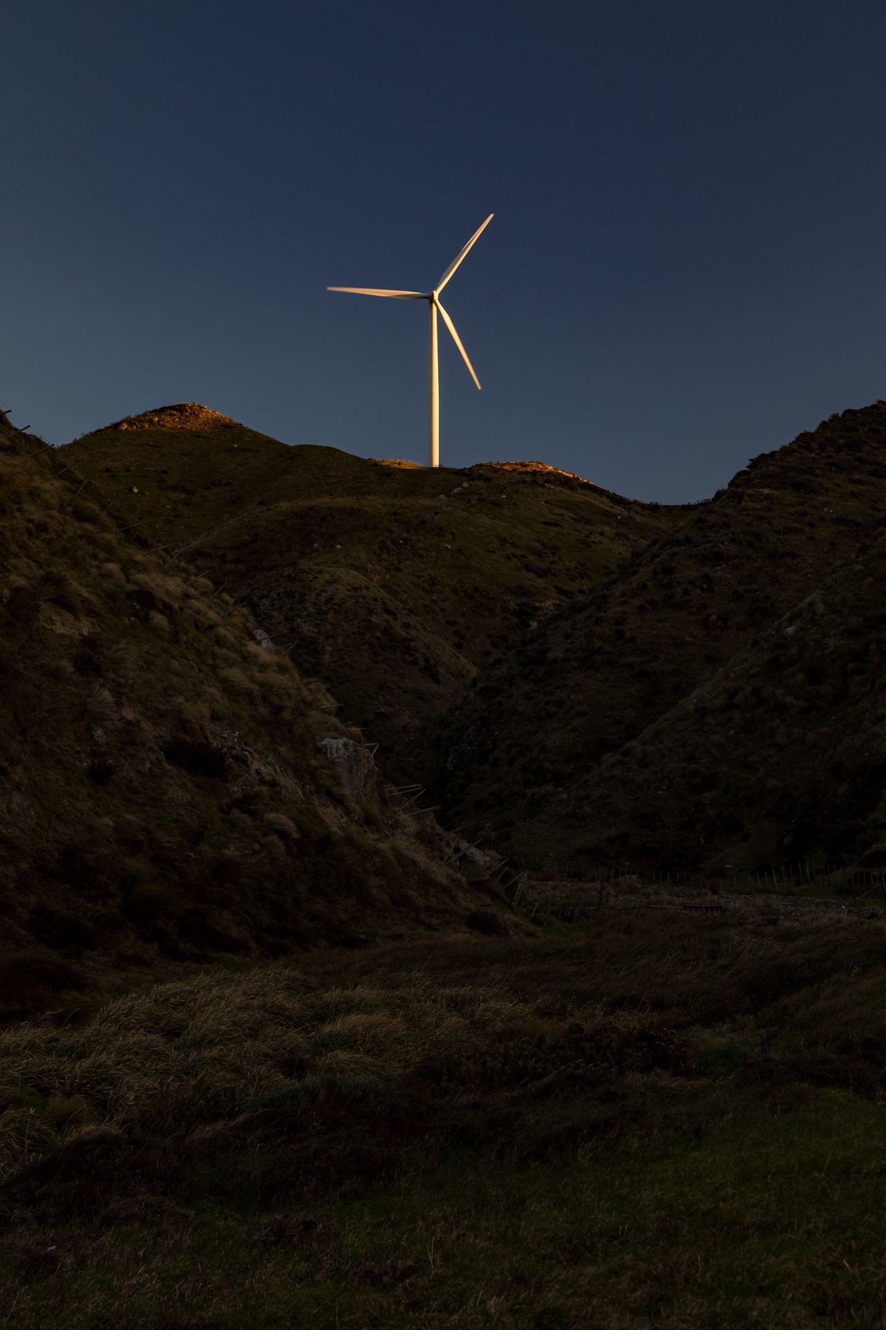 Single wind turbine lit by golden light on a dark hilltop silhouetted against a deep blue evening sky.