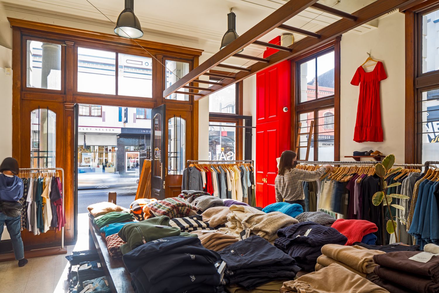 Bright clothing boutique with stacked folded sweaters, hanging garments, a red dress in the window and shoppers browsing.