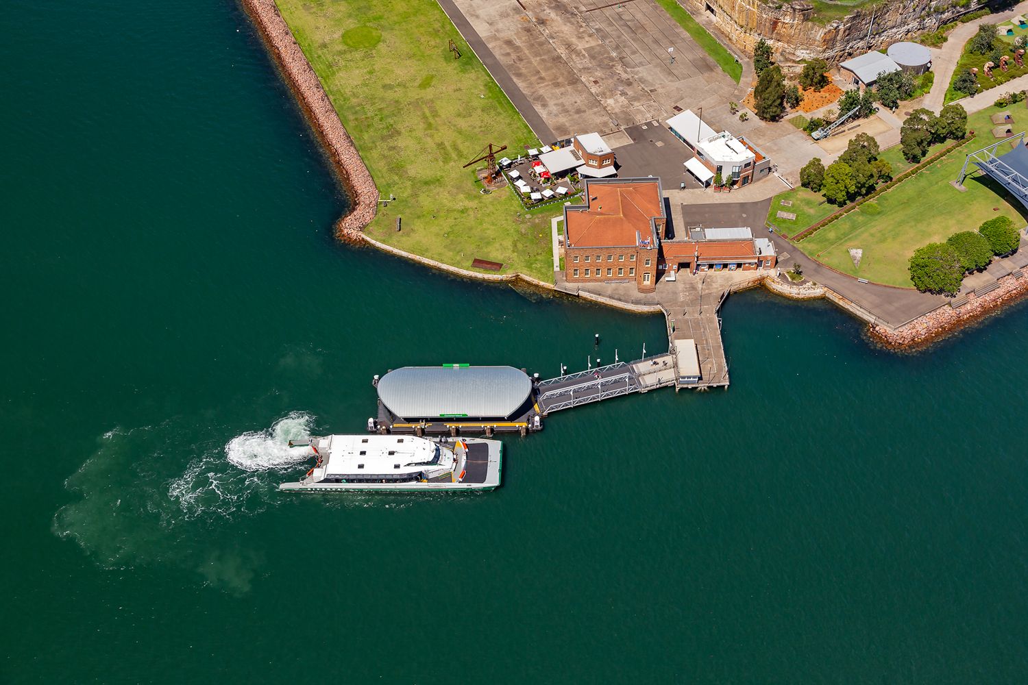 Aerial view of a ferry arriving at a canopy-covered harbour wharf near historic red brick buildings and green grassy foreshore.