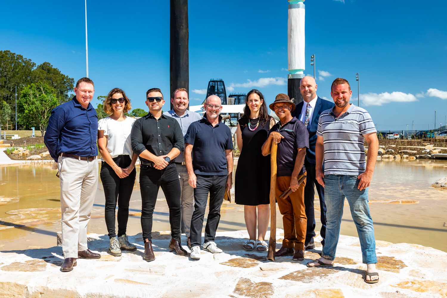 Group of ten people pose together on a sandstone waterfront plaza beside tall painted poles on a bright sunny day.