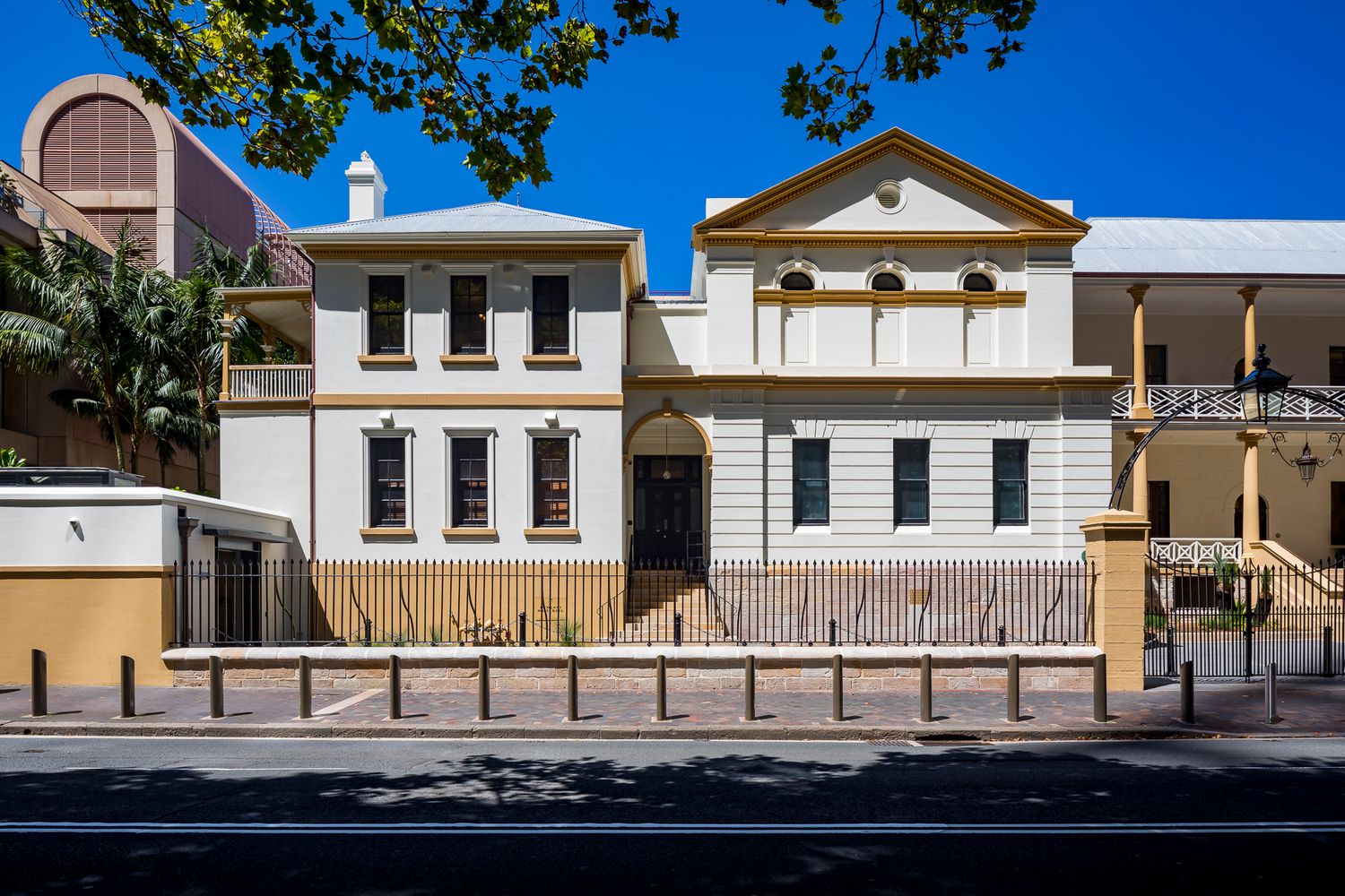 Historic two-storey cream and ochre building with pitched gable roof, arched entrance and iron fence, seen across a street under blue sky.
