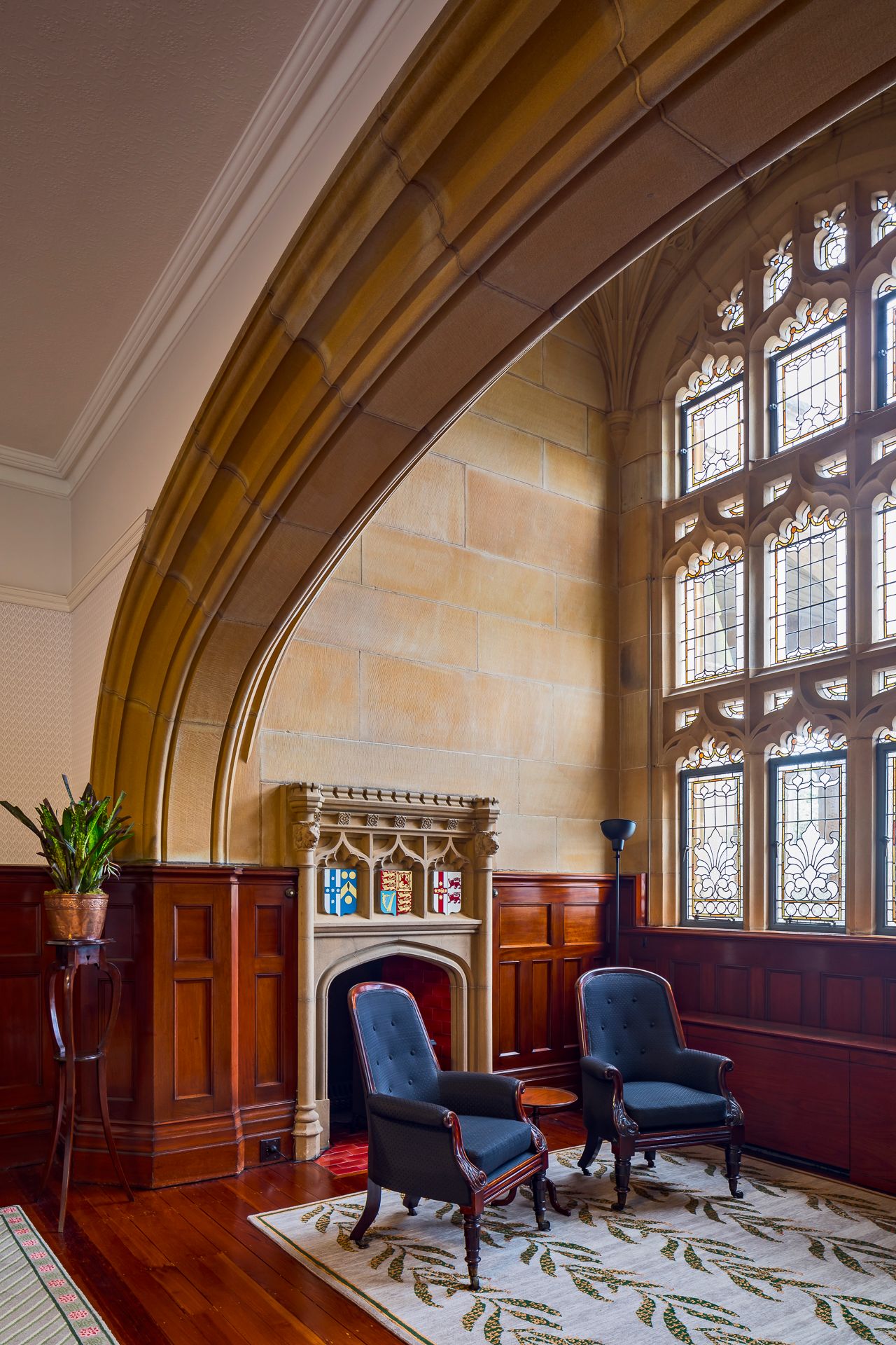 Historic room with a sandstone Gothic arch, leaded stained glass window, carved stone fireplace, blue armchairs and timber panelling.