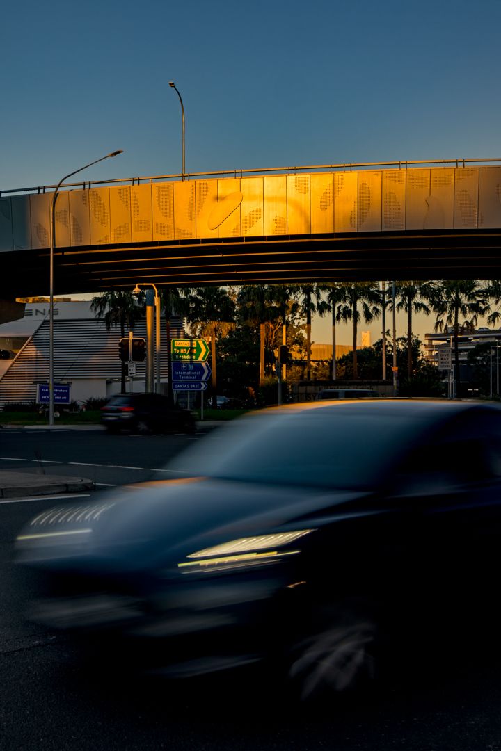 Blurred dark car passes beneath a golden-lit overpass at dusk with palm trees and road signs in the background.