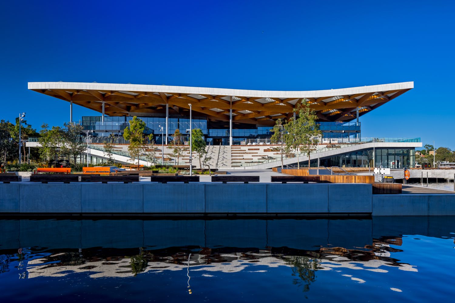 Waterfront pavilion with a wide timber-lined cantilevered roof, outdoor steps and trees reflected in calm water under a deep blue sky.