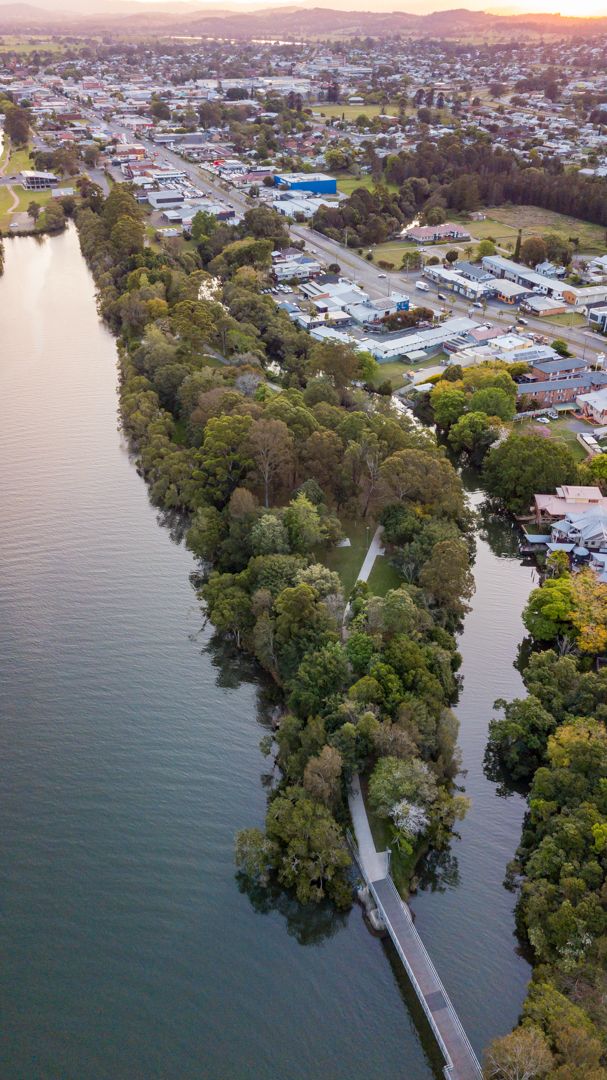 Aerial view of a tree-lined riverbank with a narrow footbridge and town buildings in the background at sunset.
