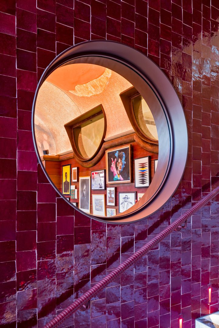 Round porthole window in glossy burgundy tiled wall revealing a warm interior with framed artwork on the walls.