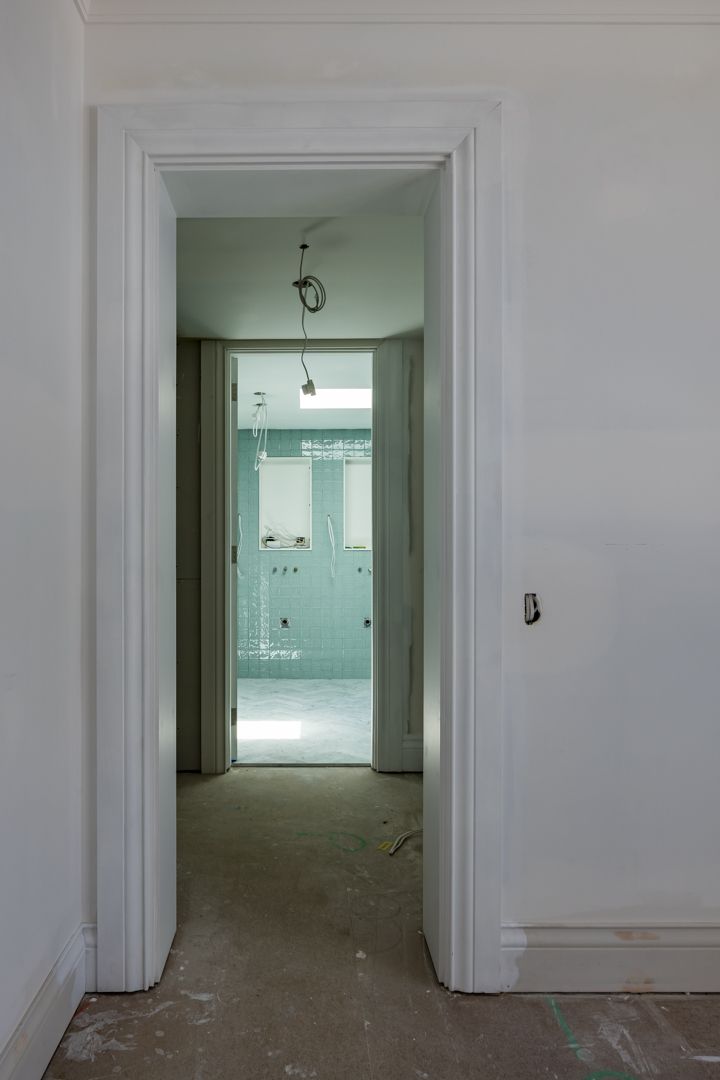 Unfinished white-walled hallway with timber trim leading to a bathroom lined with green waterproof board and exposed ceiling wiring.