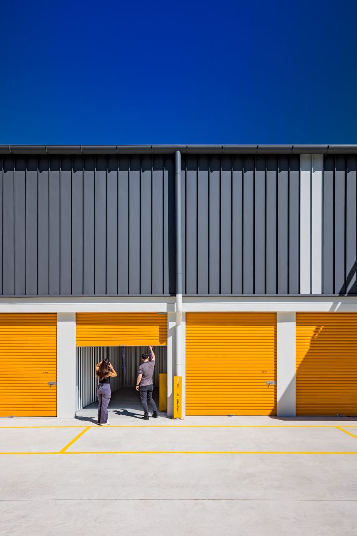 Two people stand at an open unit in a self-storage building with bright yellow roller doors and dark grey cladding under a blue sky.