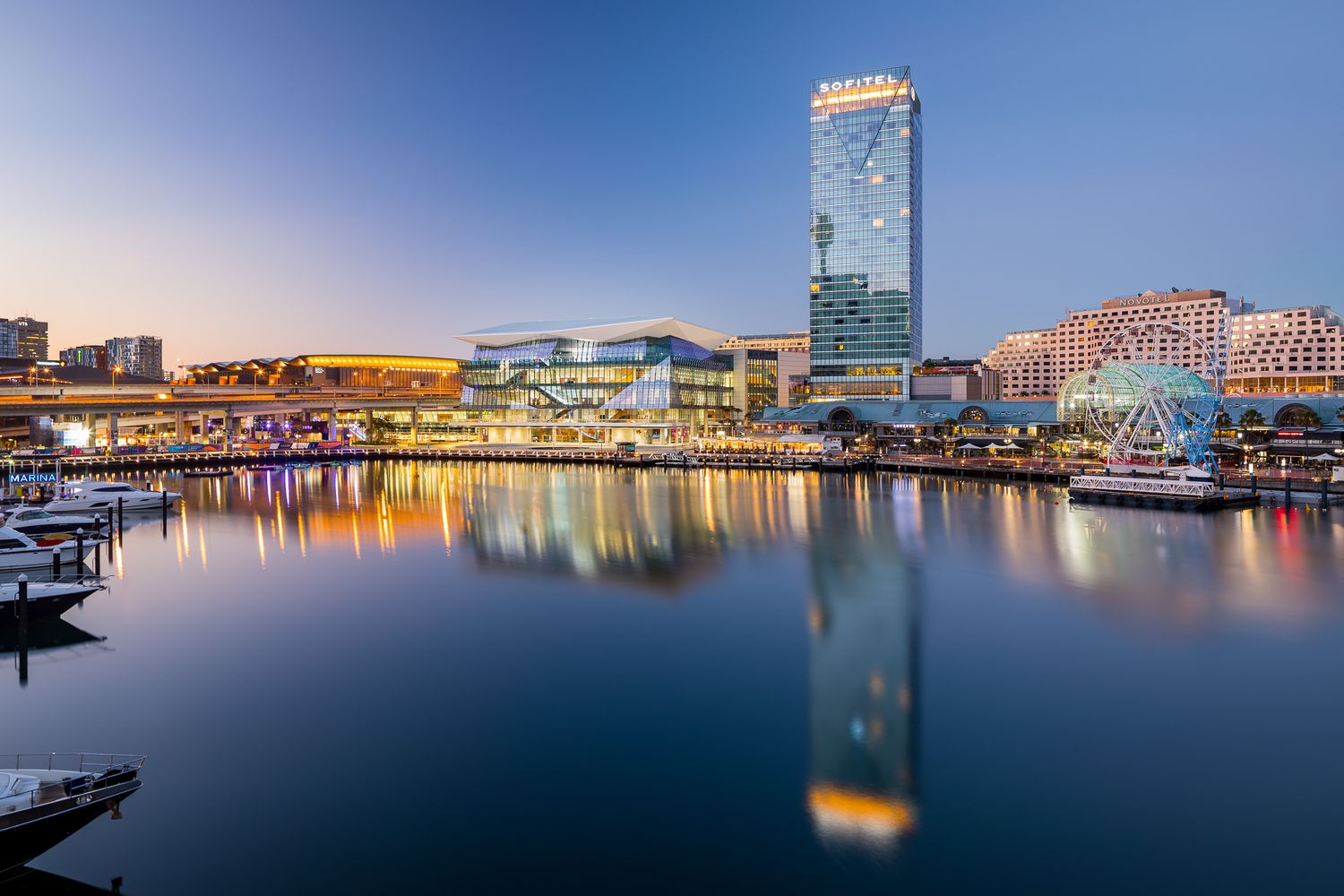 Waterfront precinct at sunset with a glass convention centre and high-rise tower reflected in the calm harbour beside moored boats.