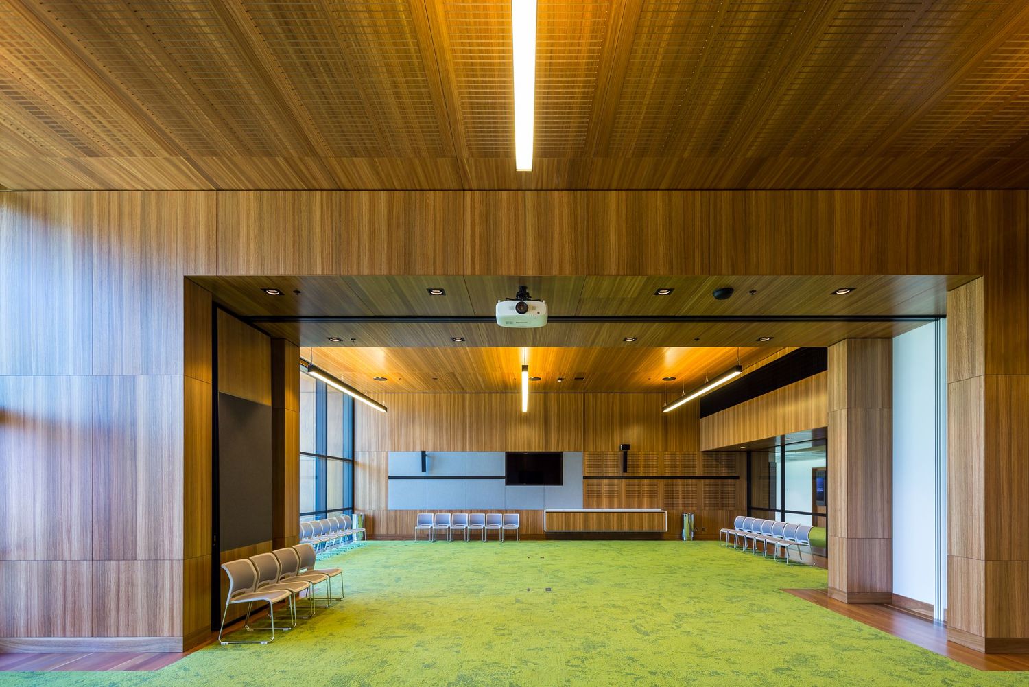 Spacious meeting room with warm timber wall and ceiling cladding, bright green carpet and built-in presentation screen.