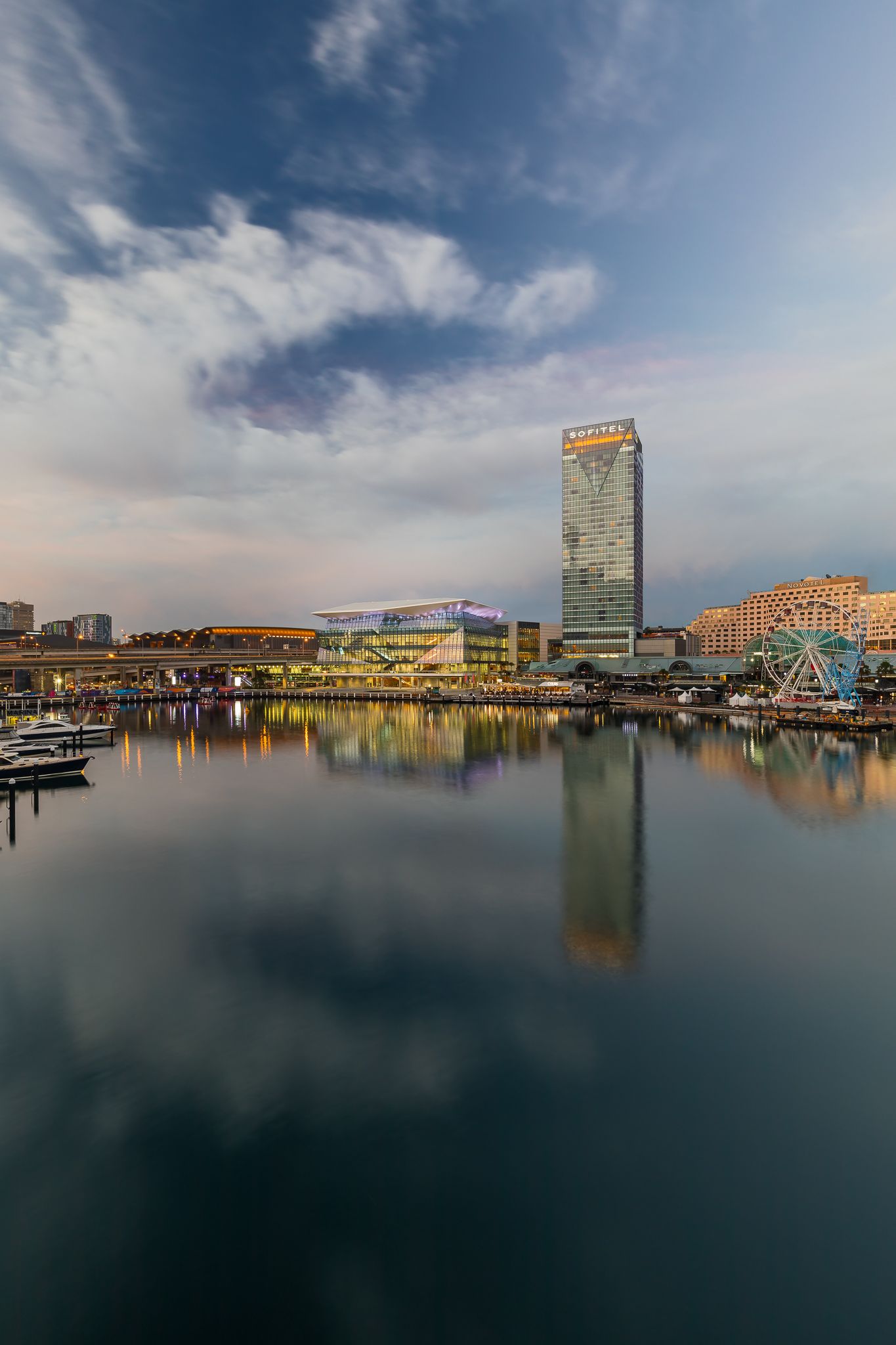 Darling Harbour skyline at dusk with a tall hotel tower and ferris wheel reflected in calm harbour water.