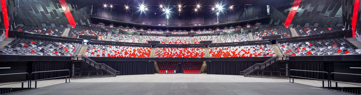 Wide panoramic view of a large indoor arena with tiered red seating, bright overhead lights and an open floor ready for events.