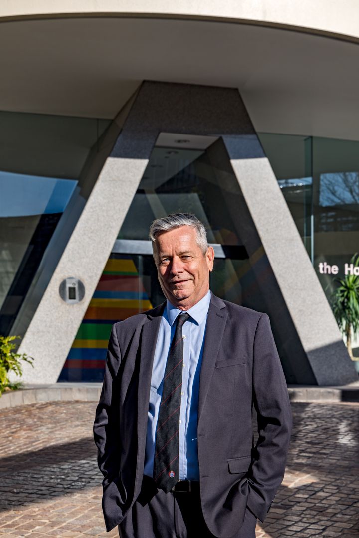 A smiling gray-haired man in a dark suit and tie stands outdoors in front of a modern building with angular concrete columns.