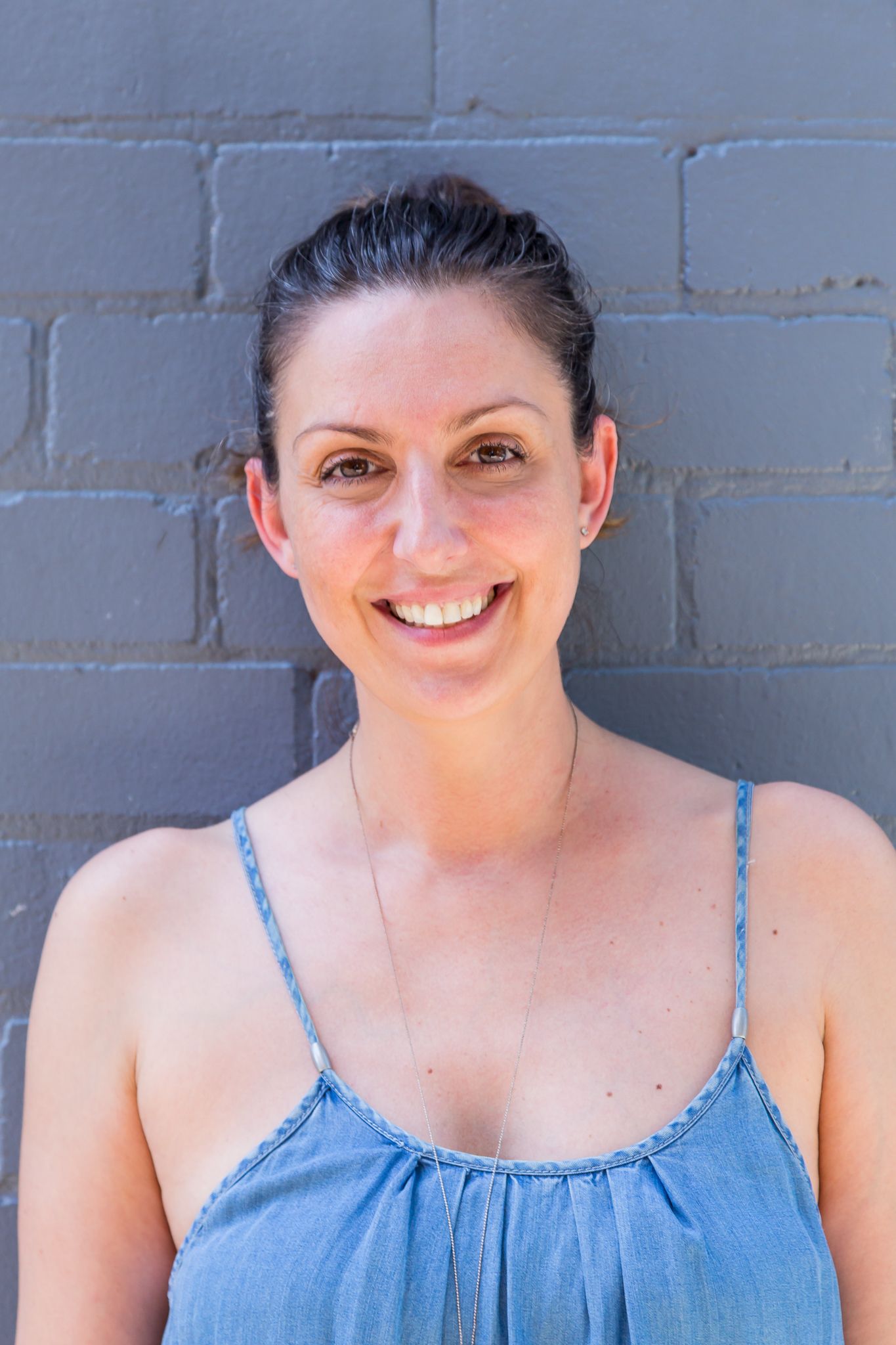 A smiling woman with her hair pulled back wears a blue sleeveless top and necklace while standing against a gray painted brick wall.