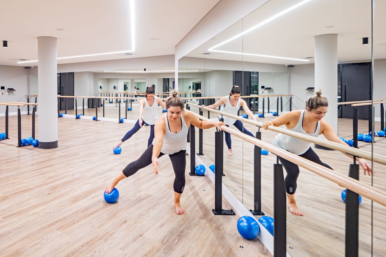 Women exercise at a bright barre fitness studio, holding the barre while balancing with small blue pilates balls under their feet.