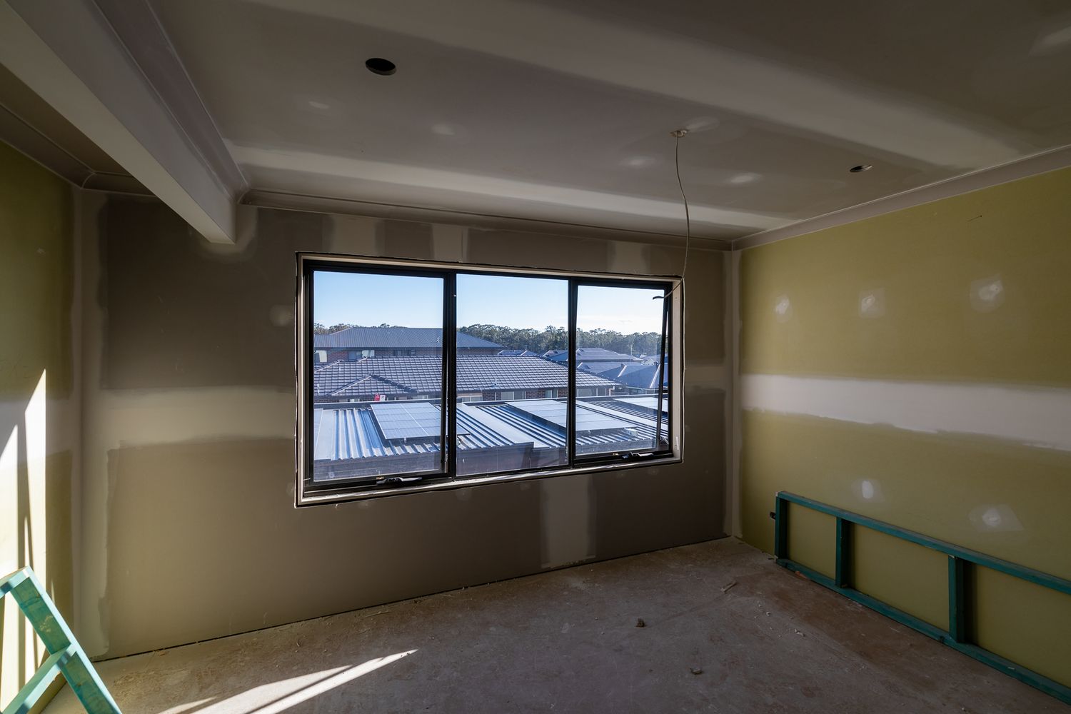 Unfinished upstairs room with plasterboard walls, taped joints and a large black-framed window overlooking neighbouring rooftops.