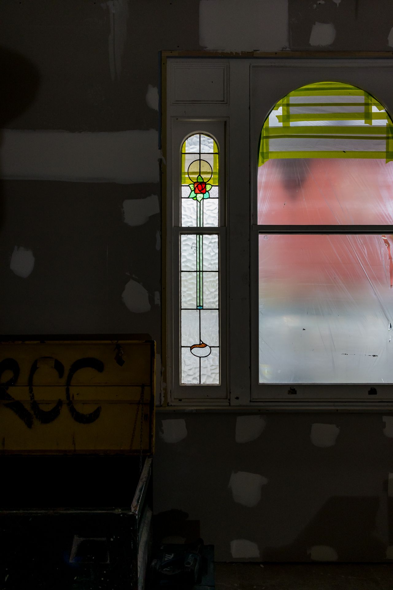 Dimly lit interior with an arched stained glass window and a tall narrow leadlight panel beside patched plasterboard walls.