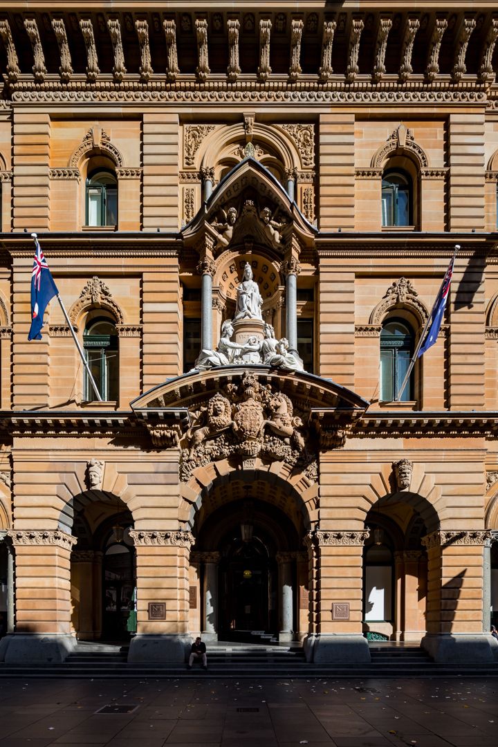 Ornate sandstone Victorian building facade with arched entrance, sculpted statues, carved coat of arms and flags flying on both sides.