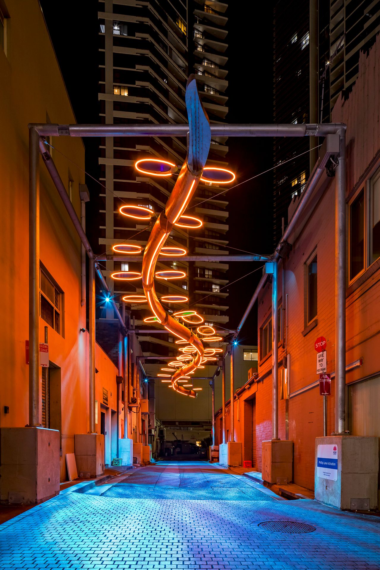 An amber neon spiral light sculpture glows above a city alley with golden walls and blue-tinted pavement.