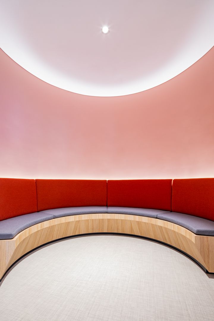 Curved built-in bench with red cushions and a timber base beneath a round pink illuminated ceiling in a modern office breakout space.