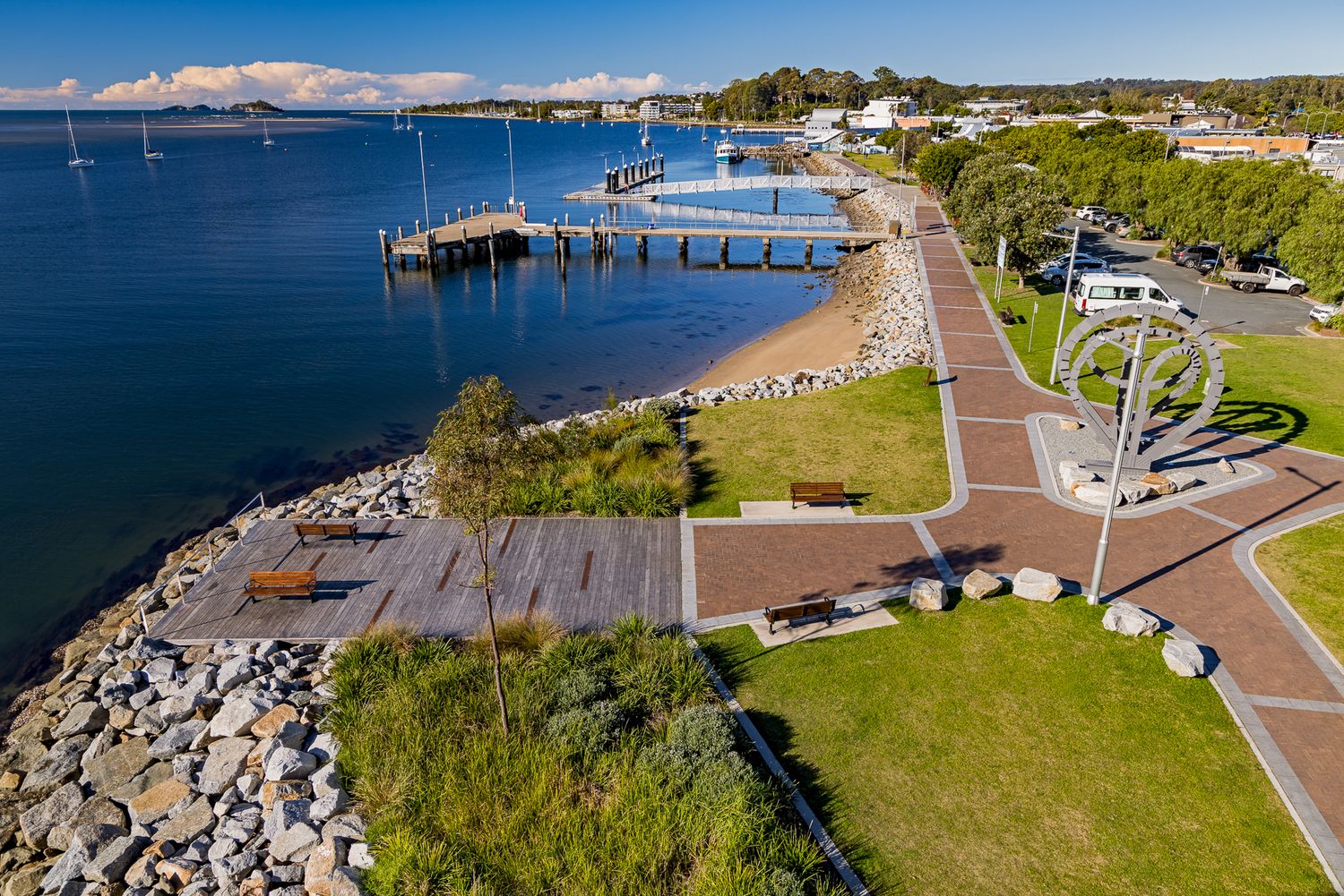 Aerial view of a waterfront park with a timber jetty, rocky shoreline, walking paths and a circular sculpture.