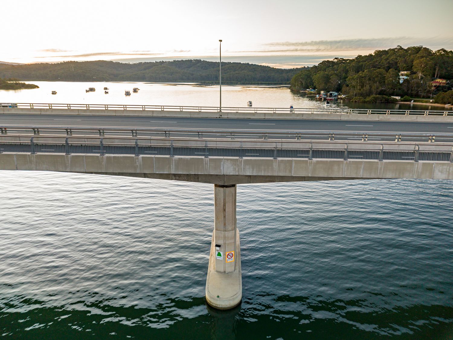 Concrete bridge pier rising from calm blue water with a waterfront bridge deck and forested hills behind.