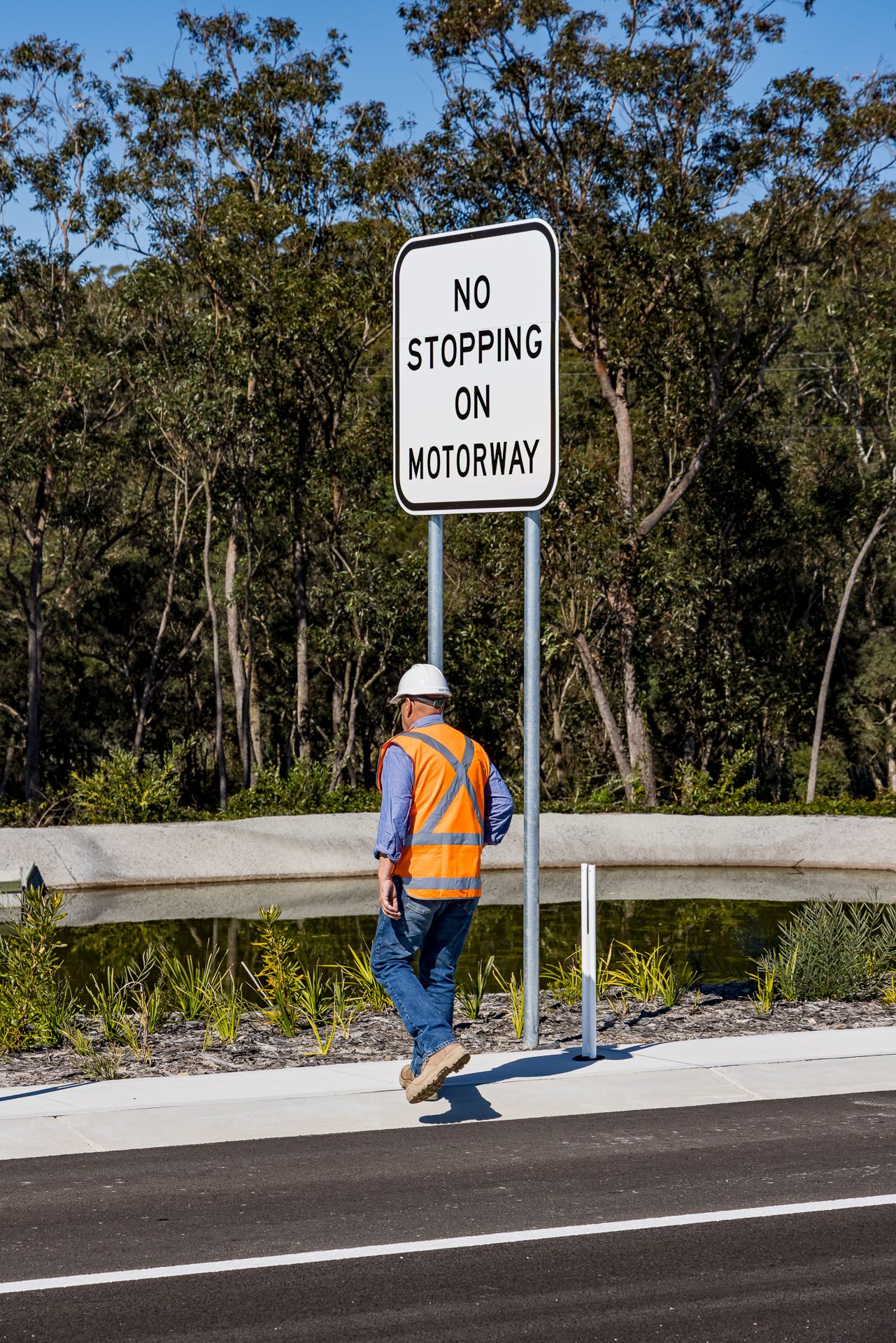 Construction worker in hard hat and orange high-visibility vest walking beside a No Stopping on Motorway sign near bushland.