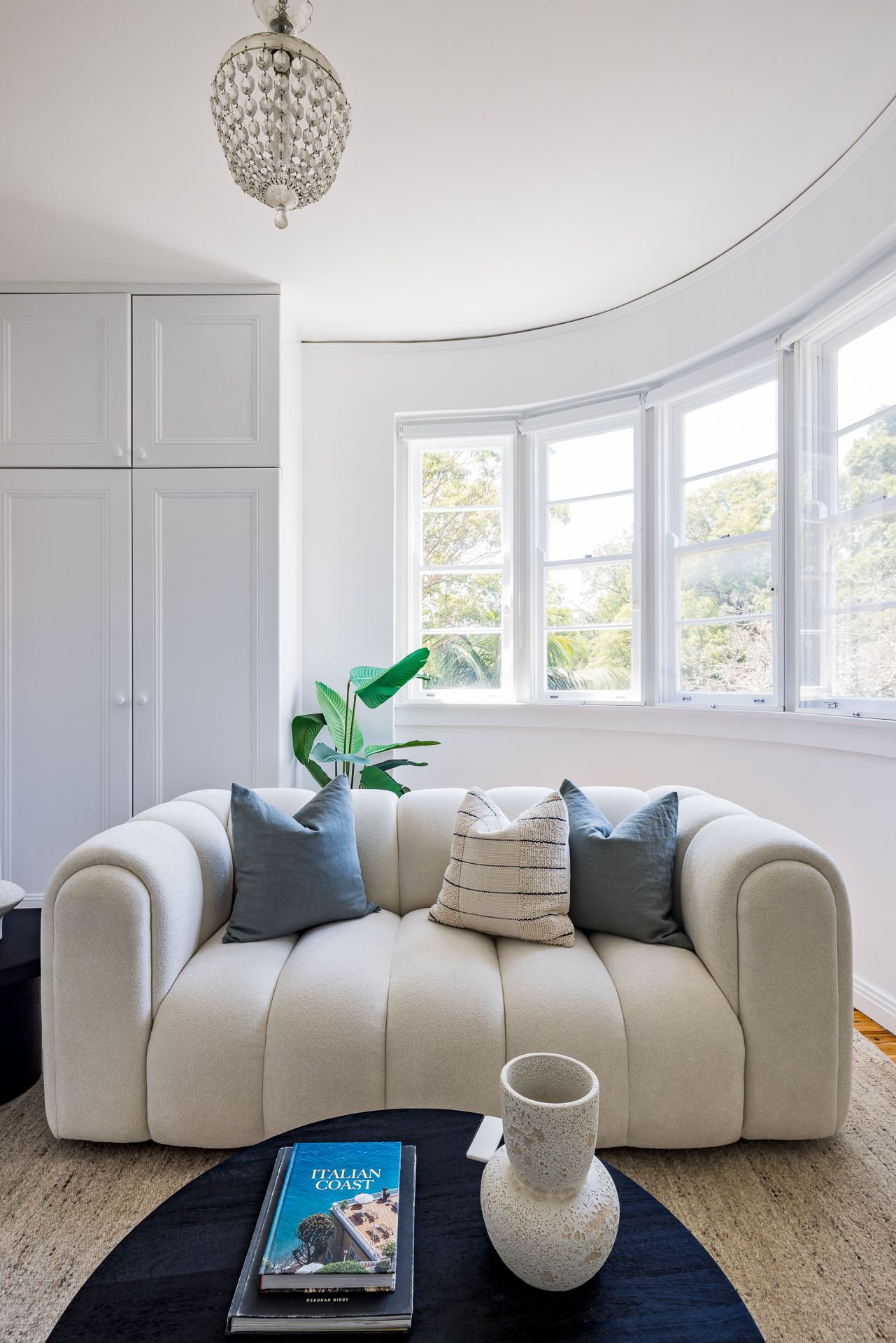 Curved sunroom with cream boucle sofa, cushions, round black coffee table, crystal chandelier and leafy window views.