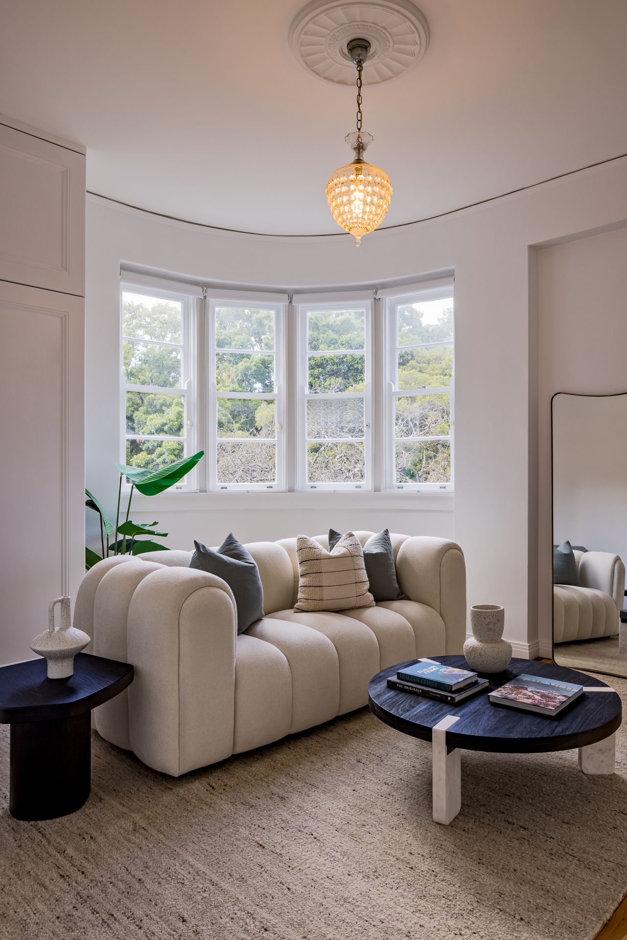 Cozy living room corner with a cream curved sofa, bay windows, crystal pendant light, round dark coffee table and woven jute rug.